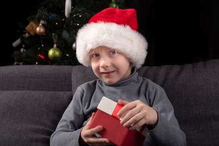 Portrait of boy in Santa hat with gift in his hands. Little child sitting on the sofa against the background of the Christmas tree.の写真素材