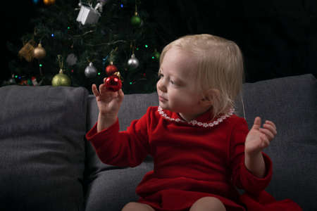 little girl sits on a sofa against the background of a Christmas tree and looks at a Christmas tree...