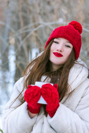 Portrait of beautiful girl in red knitted hat and mittens with cup in her hands. Outdoors. Vertical...