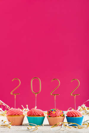 New Years background. Holiday cupcakes with numbers 2022 surrounded by New Years tinsel on pink...