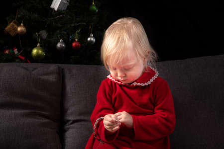 little girl sits on a sofa against the background of a Christmas tree and looks at a Christmas tree...
