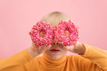 Fair-haired baby holds pink donuts near his eyes. Portrait of boy with donut on pink background