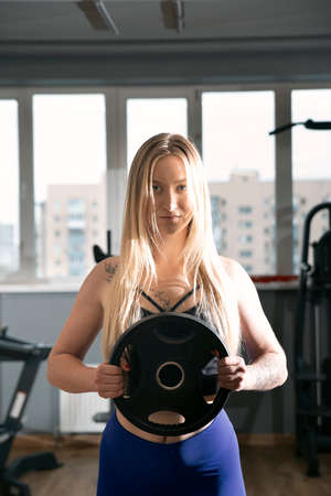 Sexy caucasian fitness model posing in the gym. Blonde girl is holding dumbbells. Sport, healthy lifestyle. Vertical frame.の写真素材