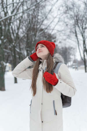 Brooding young woman in red knitted hat and mittens walks through snow-kept park. Winter dayの写真素材