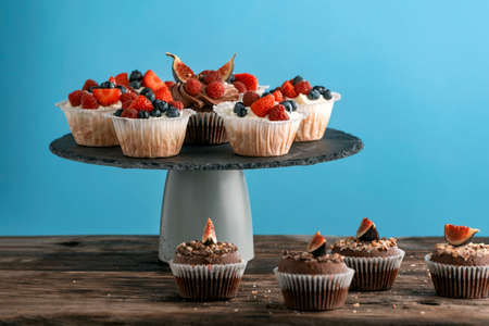 Confectionery on stand. Vanilla and chocolate muffins with fresh berries on blue background. Side...