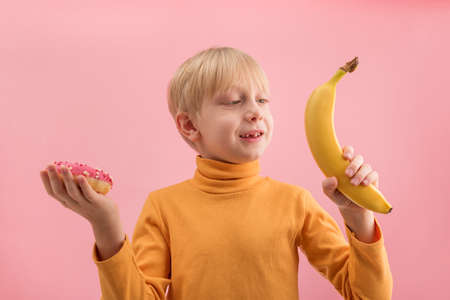 Young blond boy choosing between fruits and sweets. Child holding donut and banana. Healthy foodの写真素材