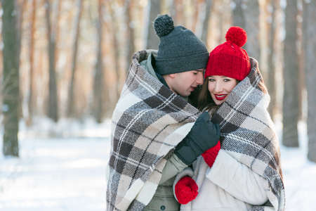 Young couple are hugging each other in a snow-covered park, wrapping themselves in a plaid. Man and woman in winter forestの写真素材
