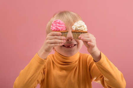 Adorable happy fair-haired boy smiling and holding yummy cupcakes in hands on pink backgroundの写真素材