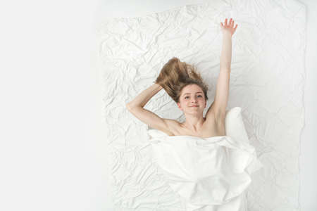 Beautiful naked woman lies in bed hands up, covered with white sheet and enjoys the weekend. Relaxing at home. White background. Top view.の写真素材