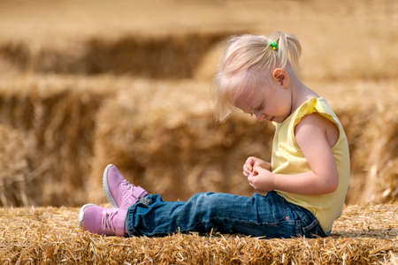 Little blonde girl sits in the hay. Child plays haystack. Childhood on the farm. Summer dayの写真素材
