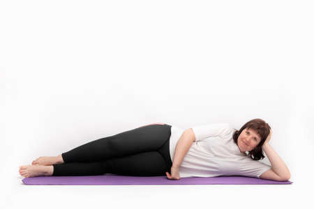 Portrait of an overweight young woman lies and looks at the camera on yoga mat on white background. Plus size model goes in for sportsの写真素材