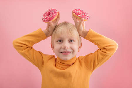 Funny child boy in an orange turtleneck with donuts over his head on pink background. Blond boy and bunsの写真素材
