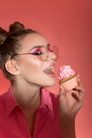 Close up portrait girl in glasses on pink background. Woman trying fruit cupcake. Pinkmood. vertical frame.の写真素材