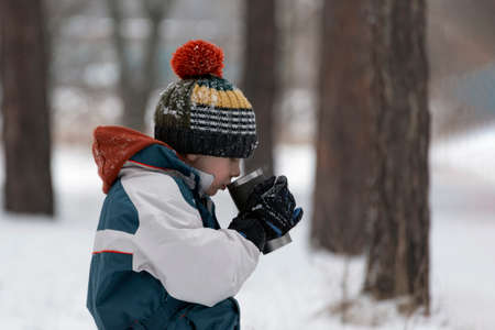 Child in knitted hat resting and drinking hot tea or cocoa from thermos on cold winter day in the forestの写真素材