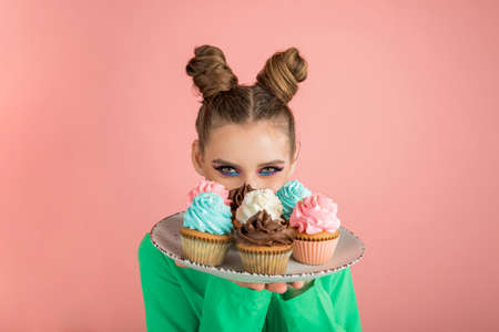 Young woman with funny buns hairstyle holds plate with colorful cupcakes. Portrait of beautiful girl with cakes on pink backgroundの写真素材