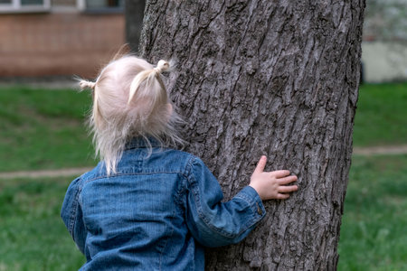 Little fair-haired girl hugging a tree in the park. Girl with two ponytails in denim jacket outside. Spring walking.の写真素材
