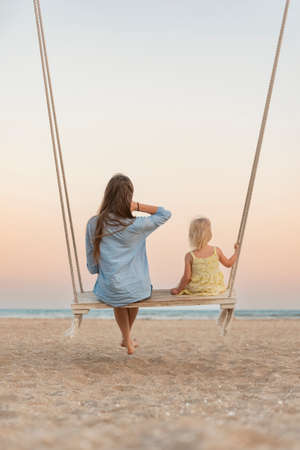Mom and little girl sit on swing and watch the stunning sunset over the sea. Magical sunrise over seaの写真素材