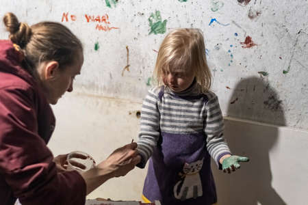 KHARKIV, UKRAINE- 3 MARCH 2022: mother and little girl paint their palms with paint to leave an iprinmt on wall. Ukrainian children in basement during war with Russiaのeditorial素材