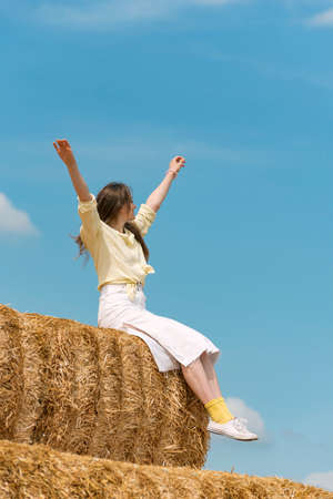 Girl in bright yellow socks sits on haystack with her hands up. blue sky. outdoor recreation countryside. harvesting.の写真素材