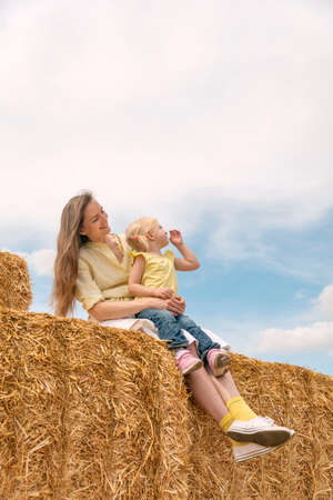Mother and child are having fun on the farm. Mother and fair-haired daughter are sitting on haystack in the field.の写真素材