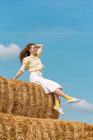 Portrait of happy young woman sitting on high haystack on blue sky background. woman resting in...