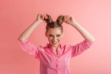 Cheerful young woman in pink shirt adjusts her hair. Girl with two bun hairstyles on pink background