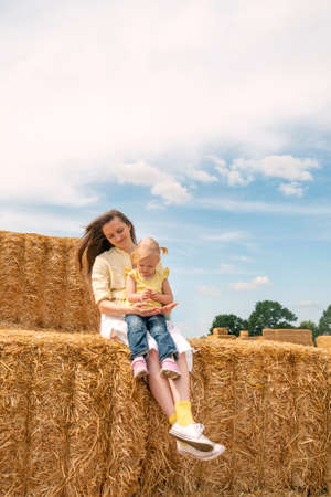 Portrait of young happy mother with little daughter in her arms sitting on haystack on farm. Hay...