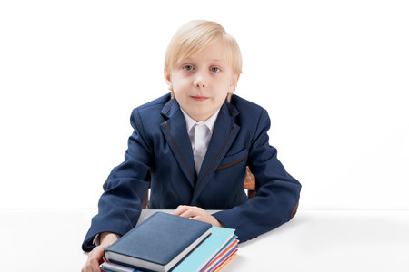 Blond boy of 7-8 years old in jacket or school uniform sits at table with stack of notebooks and...
