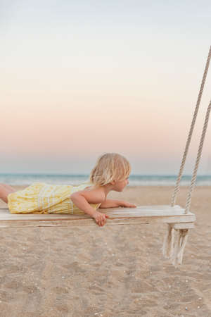 Fair-haired girl play on wooden swing on summer evening on sea background. Small female child on swing on the beachの写真素材