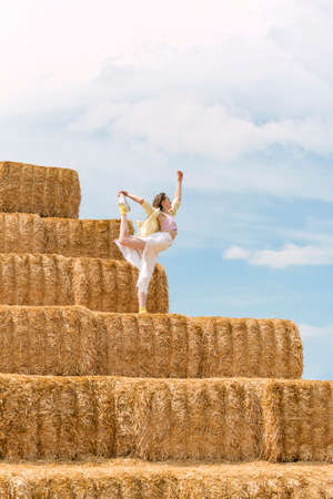 Girl dances on huge block of hay. Hay harvesting for autumn. Woman practicing yoga on haystackの写真素材