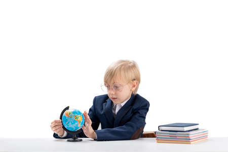 First grader boy in big glasses and school uniform sits at desk and carefully examines the globe....