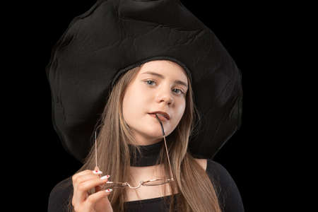 Portrait of young girl in wide-brimmed black hat with glasses in her hands. Studio portrait of lady on black backgroundの写真素材