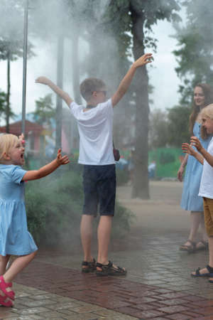 Children fool around in the park rejoicing in breeze. Water sprayer for people on hot day. Walk in...
