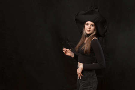 Young woman in form of witch wears black clothes, pointed hat and holds glasses. copyspace. Isolation on black background.の写真素材