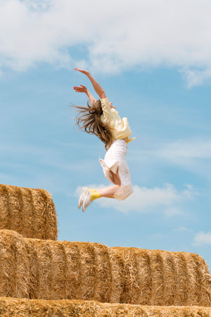 Cheerful young woman jumps from high haystack. Happy girl has fun on the farm on blue sky backgroundの写真素材
