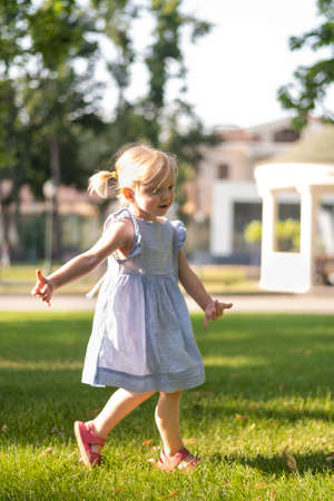 Little blonde girl in blue dress walking in the park. Three-year-old child in sundress with two tails on trees backgroundの写真素材