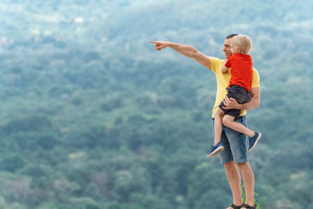 Father holds his son in his arms and points his finger somewhere in the distance. Father and son on hill in the mountainsの写真素材