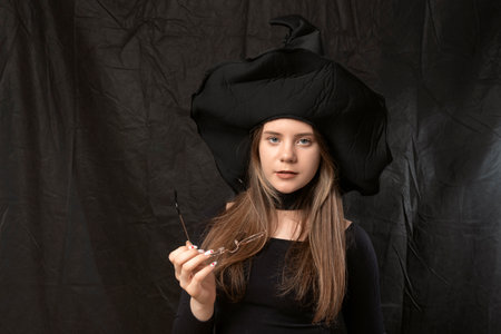 Portrait of young girl in pointed witch hat with glasses in her hands on black background. halloween...