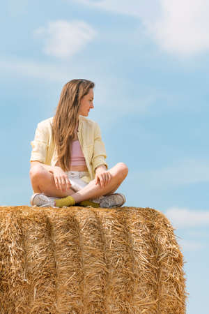 Girl with long blond hair sits on haystack cross-legged. Portrait of woman in the hay on farmの写真素材