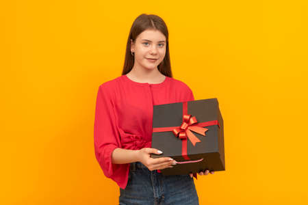 Young girl holds large black box with red bow on bright yellow background. Young woman with gift in...