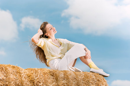 Female model posing in the field after harvesting by the haystack. Young woman sitting on big straw stacksの写真素材