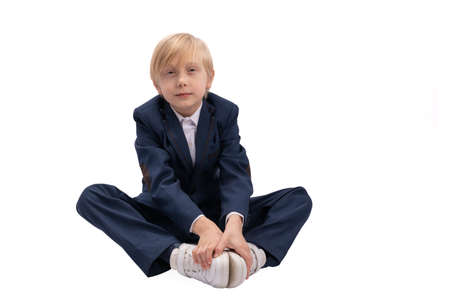 Blond boy in school uniform sits with his hands on sneakers. Portrait of student of the 1st or 2nd grade in isolation on white backgroundの写真素材