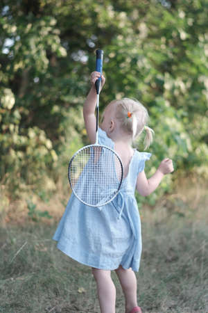 Blonde three years old girl with two tails and summerdress plays badminton. Child in blue dress holds racket and shuttlecockの写真素材