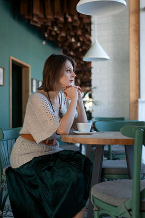 Brooding young woman sits at table in an empty cafe with cup of coffee. Lonely lady in the cafeteria