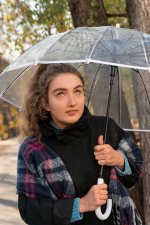 Portrait of young woman looking up from under transparent umbrella. Girl with blue eyes and brown...