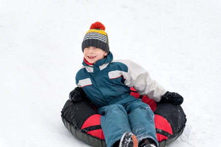 Happy boy in winter jumpsuit and hat with bubo rides on tubing in snow. Merry winter fun.の写真素材