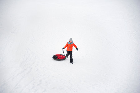 Boy in orange jacket runs through snow drags inflatable sled. Young woman on snow background in...