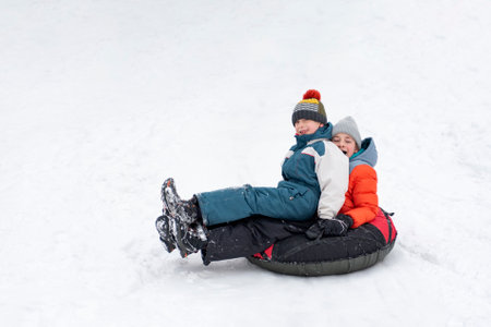 Children in winter jumpsuits and knitted hats slide down slope on inflatable sled. Boys have fun in winter outsideの写真素材
