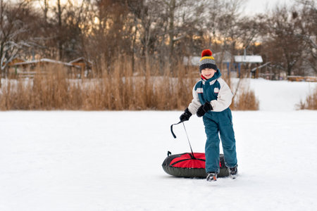 Cheerful boy in winter jumpsuit and bright hat runs through snow with tubing. Child on winter vacation is having fun.の写真素材