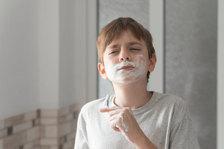 Little boy smears shaving foam on face looks in mirror with serious look. Morning routine in the bathroom.の写真素材